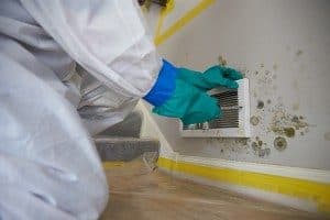 Person in a white mold removal suit with yellow gloves, removing mold from a wall near an A/C vent